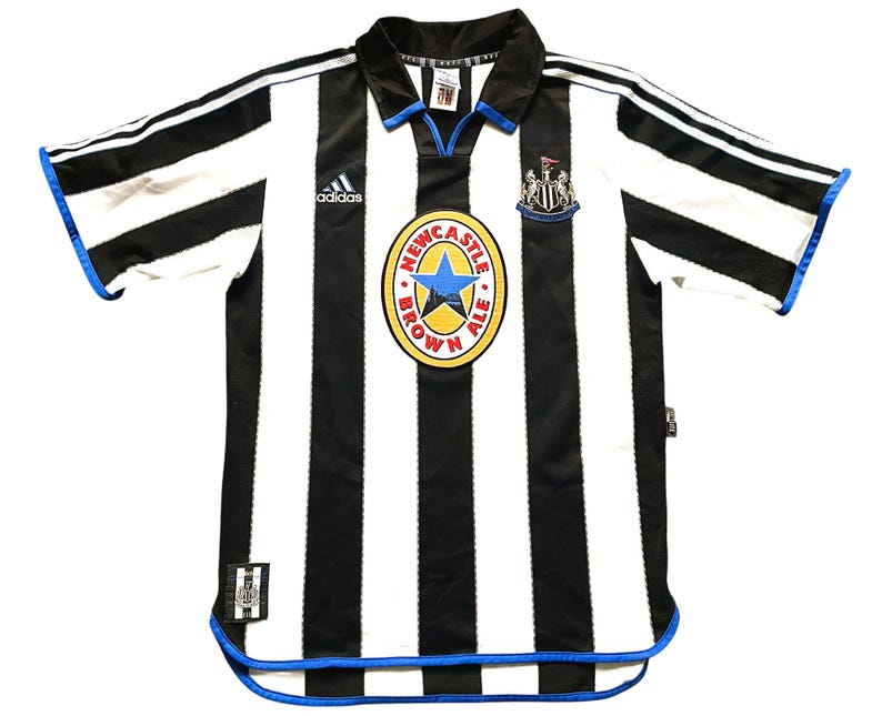 May include: Black and white striped soccer jersey with blue accents. Features the Adidas logo, a Newcastle United crest, and a circular emblem with text and a blue star. The jersey has short sleeves.