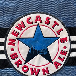 May include: A white circle patch with a red border and a blue star in the center. The star has a cityscape silhouette inside. The text "NEWCASTLE BROWN ALE" is written around the star in a red, white, and black font.