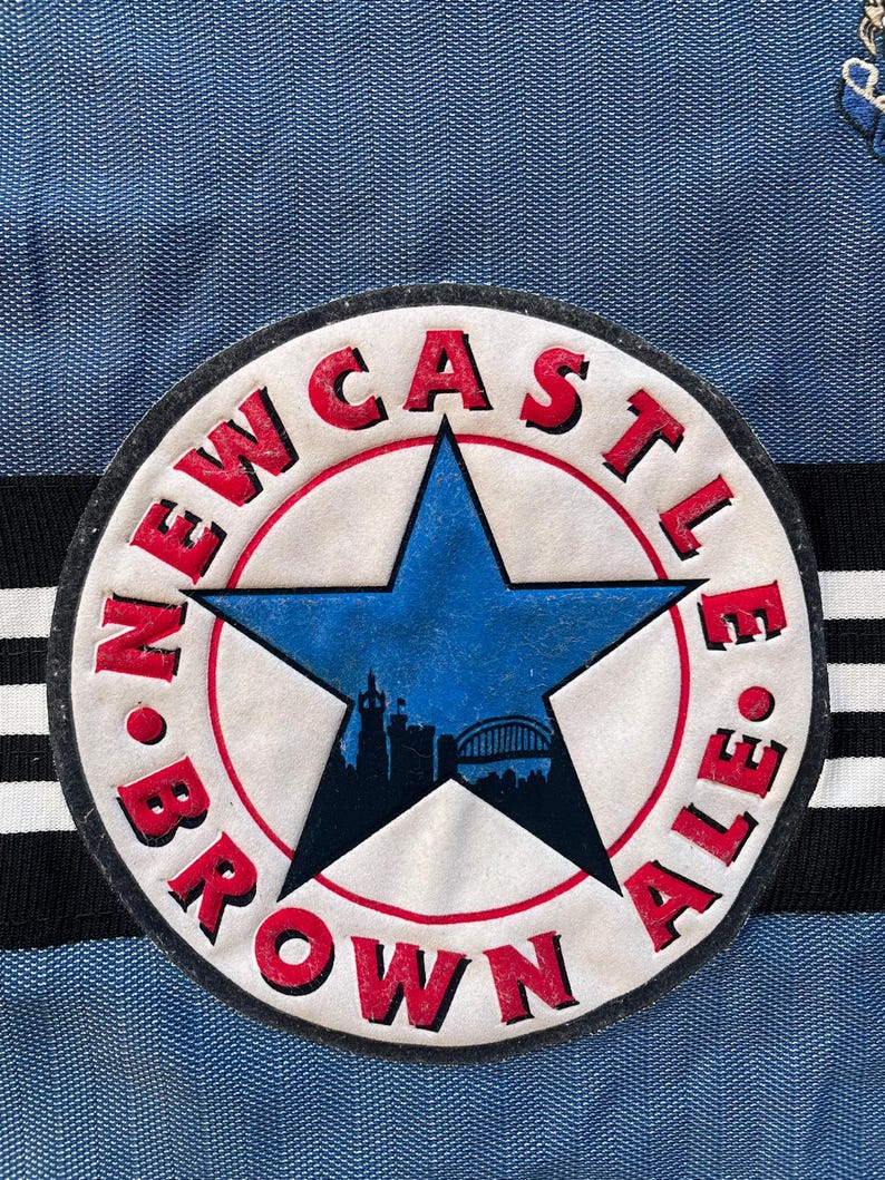 May include: A white circle patch with a red border and a blue star in the center. The star has a cityscape silhouette inside. The text "NEWCASTLE BROWN ALE" is written around the star in a red, white, and black font.