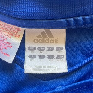 May include: A blue Adidas t-shirt with a white tag that says "Made in Tunisia, Fabrique en Tunisie". The tag also has size information in UK, USA, F, and D sizes. The sizes are 152, 12, 150, and M/M.