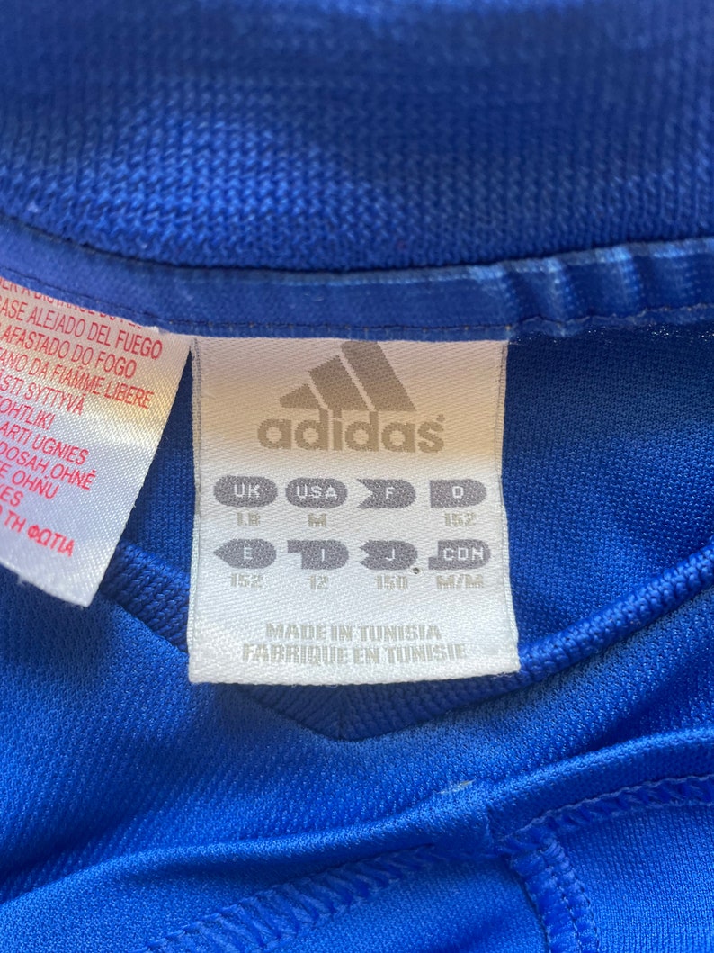 May include: A blue Adidas t-shirt with a white tag that says "Made in Tunisia, Fabrique en Tunisie". The tag also has size information in UK, USA, F, and D sizes. The sizes are 152, 12, 150, and M/M.