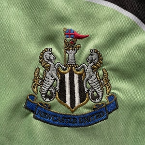 May include: Embroidered crest of Newcastle United football club on a light green fabric. The crest features a shield with black and white stripes, flanked by seahorses, and the words "Newcastle United" on a blue banner.