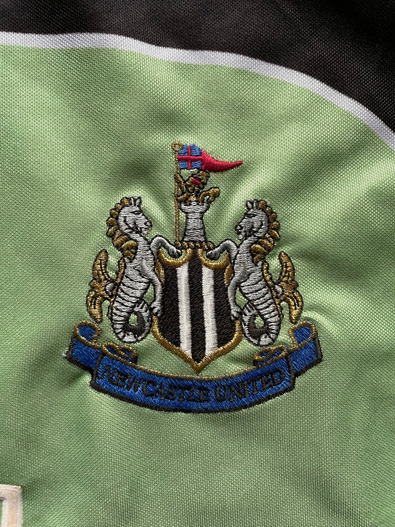 May include: Embroidered crest of Newcastle United football club on a light green fabric. The crest features a shield with black and white stripes, flanked by seahorses, and the words "Newcastle United" on a blue banner.