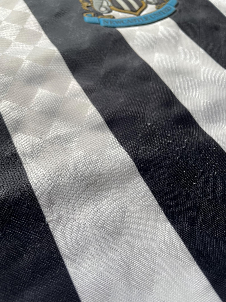 Op de afbeelding: Close-up van een vintage Newcastle United voetbalshirt. Het shirt heeft zwart-witte verticale strepen, met een ruitpatroon op de witte strepen. Het teamembleem is zichtbaar bovenaan, met "Newcastle United" op een blauwe banner.