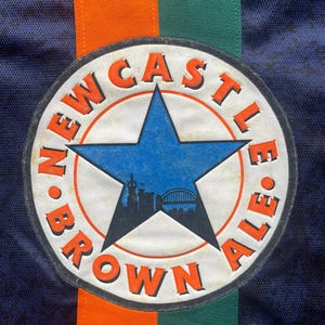 May include: Close-up of a navy blue fabric with a circular patch. The patch features a blue star with a cityscape silhouette, surrounded by the words "NEWCASTLE BROWN ALE" in orange. Vertical orange and green stripes are visible.