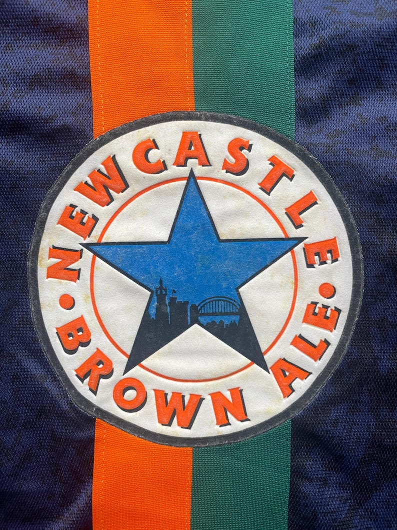 May include: Close-up of a navy blue fabric with a circular patch. The patch features a blue star with a cityscape silhouette, surrounded by the words "NEWCASTLE BROWN ALE" in orange. Vertical orange and green stripes are visible.