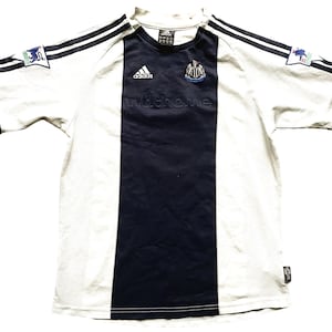 Puede incluir: Camiseta de f&uacute;tbol blanca y negra con una franja vertical negra en el centro. La camiseta tiene el logotipo de Adidas en el lado derecho del pecho y el escudo del Newcastle United en el lado izquierdo del pecho. El texto "nufc.com" est&aacute; impreso debajo del escudo.