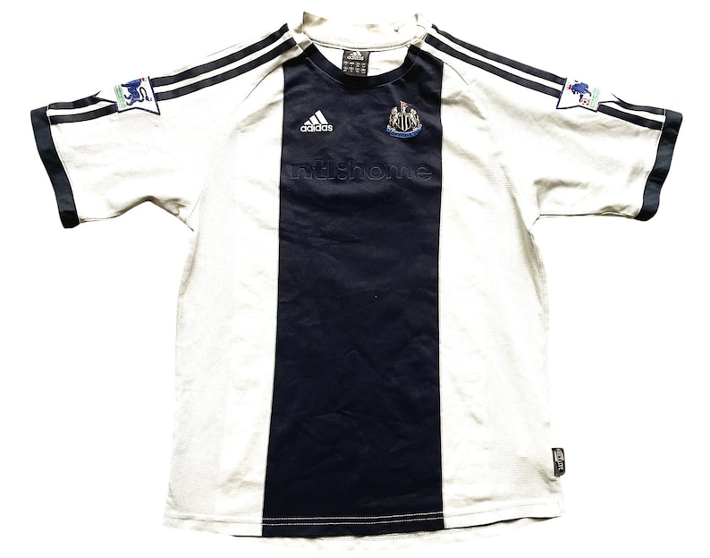 Puede incluir: Camiseta de f&uacute;tbol blanca y negra con una franja vertical negra en el centro. La camiseta tiene el logotipo de Adidas en el lado derecho del pecho y el escudo del Newcastle United en el lado izquierdo del pecho. El texto "nufc.com" est&aacute; impreso debajo del escudo.