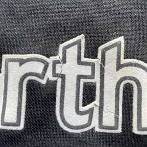 以下が含まれることがあります： 黒い衣服のクローズアップで、白い文字で「rth」と印刷されています。