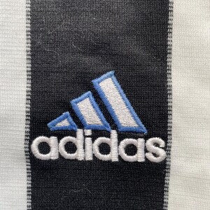 Puede incluir: Una camiseta de f&uacute;tbol a rayas blancas y negras con un logotipo de Adidas azul y blanco y la palabra "adidas" bordada en blanco.
