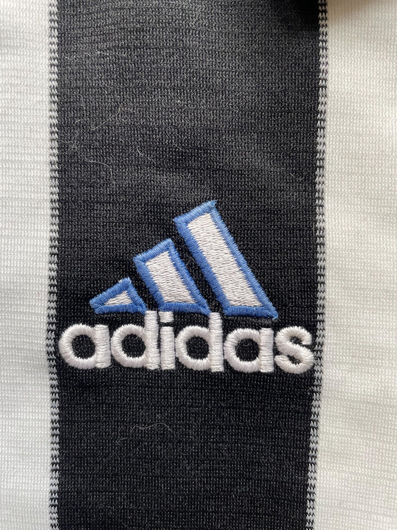 Puede incluir: Una camiseta de f&uacute;tbol a rayas blancas y negras con un logotipo de Adidas azul y blanco y la palabra "adidas" bordada en blanco.