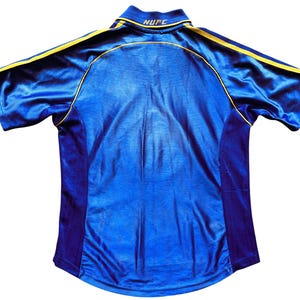 K&ouml;nnte beinhalten: Ein blaues Fu&szlig;balltrikot mit gelben und violetten Akzenten. Das Trikot tr&auml;gt den Text "NUFC" auf der R&uuml;ckseite.