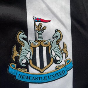 May include: Close-up of a black and white striped fabric with the Newcastle United crest. The crest features two seahorses, a shield with vertical stripes, and a banner that reads "NEWCASTLE UNITED" in blue.