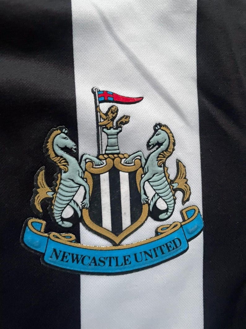 May include: Close-up of a black and white striped fabric with the Newcastle United crest. The crest features two seahorses, a shield with vertical stripes, and a banner that reads "NEWCASTLE UNITED" in blue.