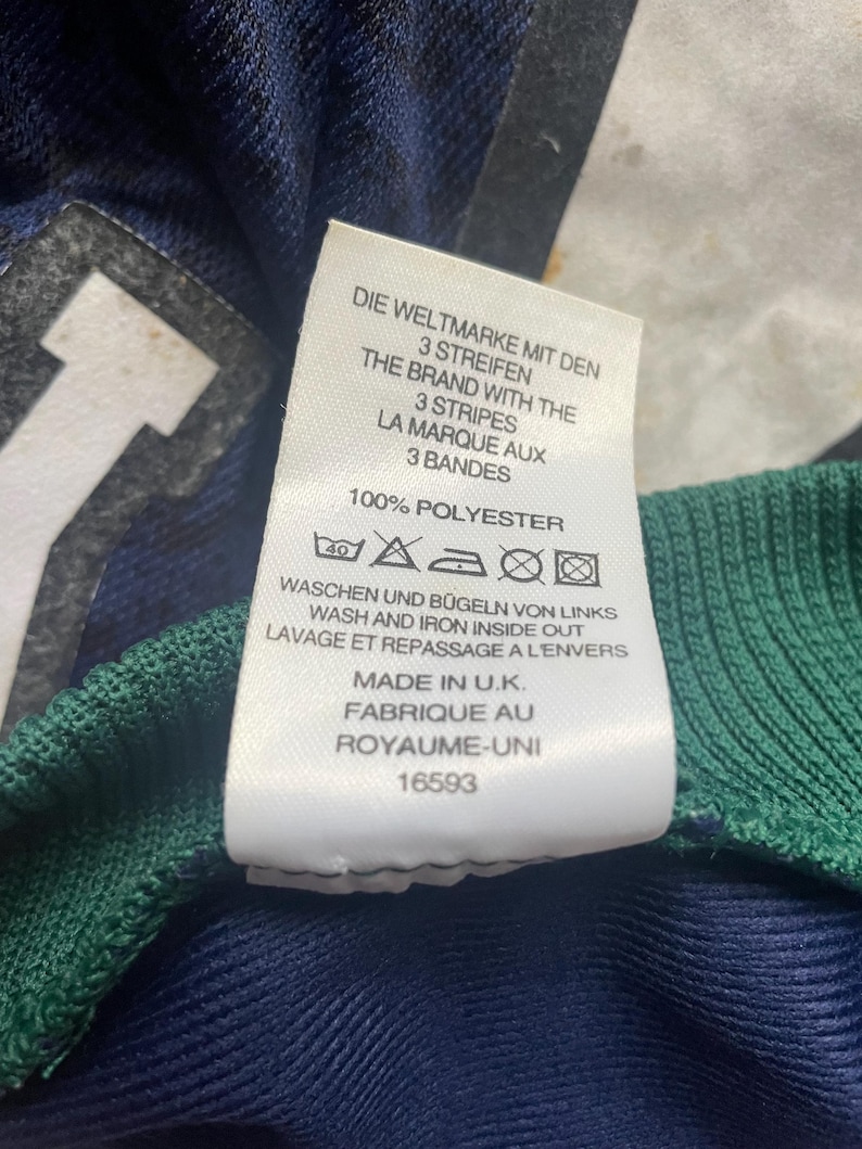 May include: Close-up of a tag with text in multiple languages, including English, German, and French. The tag reads "THE BRAND WITH THE 3 STRIPES" and "100% POLYESTER." The tag is attached to a navy blue and green garment.