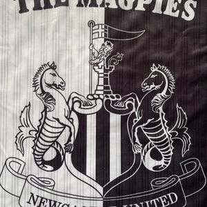 May include: A black and white striped fabric featuring the words "THE MAGPIES". The Newcastle United crest is displayed, with two seahorses, a flag, and the words "NEWCASTLE UNITED" on a ribbon.