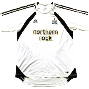 Pode incluir: Camisola de futebol branca com detalhes em preto e o texto "northern rock" em dourado. A camisola apresenta o log&oacute;tipo da Adidas e o emblema do Newcastle United. Listras pretas percorrem as mangas.