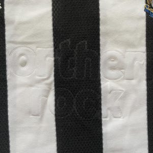 May include: Close-up of a black and white striped football shirt. The words "northern rock" are embossed on the white stripes. The Adidas logo is visible, along with a team crest.