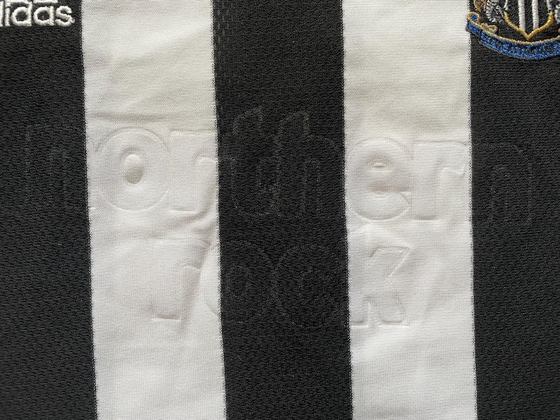 May include: Close-up of a black and white striped football shirt. The words "northern rock" are embossed on the white stripes. The Adidas logo is visible, along with a team crest.