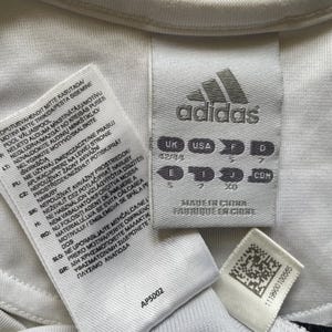 Pode incluir: Close-up de etiquetas de roupa Adidas brancas com texto em v&aacute;rios idiomas. O log&oacute;tipo da Adidas &eacute; vis&iacute;vel, juntamente com informa&ccedil;&otilde;es de tamanho e o texto "Made in China". Uma pequena etiqueta com um c&oacute;digo de barras tamb&eacute;m est&aacute; presente.