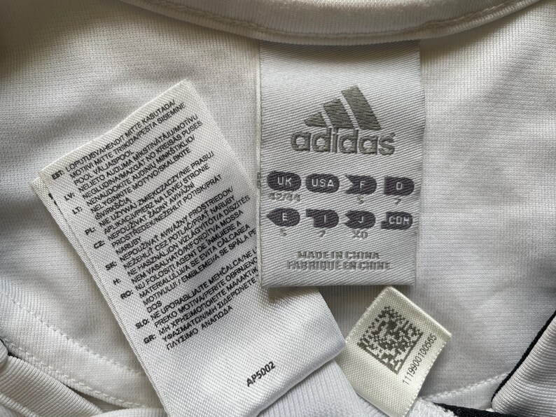 Pode incluir: Close-up de etiquetas de roupa Adidas brancas com texto em v&aacute;rios idiomas. O log&oacute;tipo da Adidas &eacute; vis&iacute;vel, juntamente com informa&ccedil;&otilde;es de tamanho e o texto "Made in China". Uma pequena etiqueta com um c&oacute;digo de barras tamb&eacute;m est&aacute; presente.