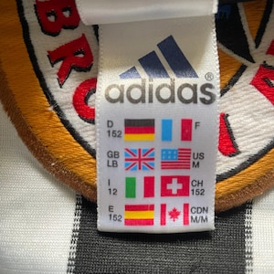 May include: Close-up of an Adidas tag with the Adidas logo and various country flags. The tag includes size information and country codes. The background is a white and black striped fabric.