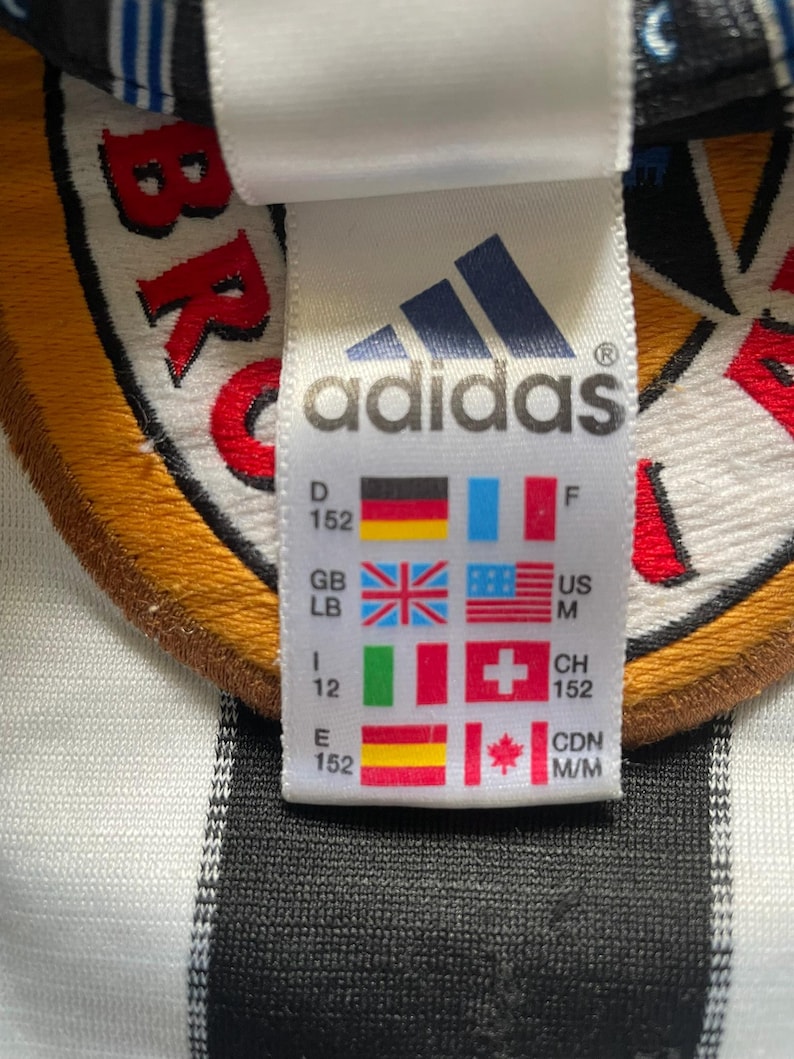 May include: Close-up of an Adidas tag with the Adidas logo and various country flags. The tag includes size information and country codes. The background is a white and black striped fabric.