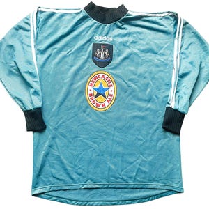 May include: A teal blue long-sleeved soccer jersey with white Adidas stripes on the sleeves. The jersey has a Newcastle Brown Ale logo on the front, along with the Newcastle United crest and the Adidas logo.