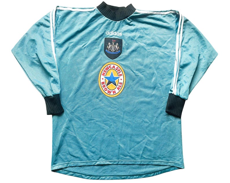 May include: A teal blue long-sleeved soccer jersey with white Adidas stripes on the sleeves. The jersey has a Newcastle Brown Ale logo on the front, along with the Newcastle United crest and the Adidas logo.
