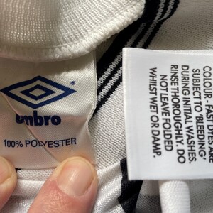 Op de afbeelding: Detail van een wit en zwart gestreepte stof met twee labels. E&eacute;n label toont het Umbro-logo en de tekst "100% POLYESTER". Het andere label geeft wasvoorschriften voor kleurechte kleurstoffen.