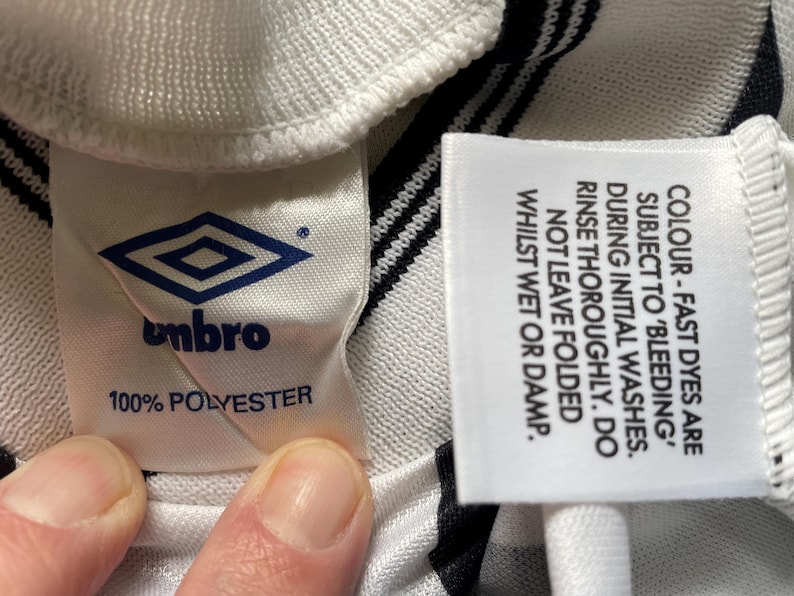 Op de afbeelding: Detail van een wit en zwart gestreepte stof met twee labels. E&eacute;n label toont het Umbro-logo en de tekst "100% POLYESTER". Het andere label geeft wasvoorschriften voor kleurechte kleurstoffen.