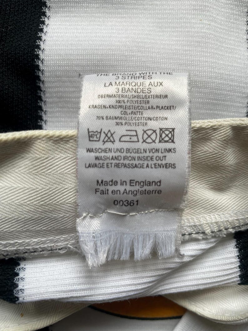May include: Close-up of a white and black striped fabric with a tag attached. The tag includes text in English, French, and German, with washing instructions and the phrase "Made in England". The fabric is likely part of a garment.