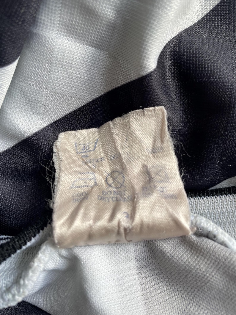 May include: A white tag with black text and symbols. The tag has instructions for washing and drying the garment. The text includes "Machine Wash", "Do Not Bleach", "Tumble Dry Low", and "Iron Low".