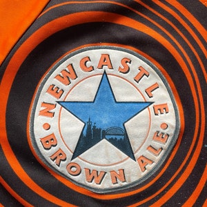 May include: A black and orange circular patch with the text "NEWCASTLE BROWN ALE" in a white circle with a blue star and a cityscape inside the star.