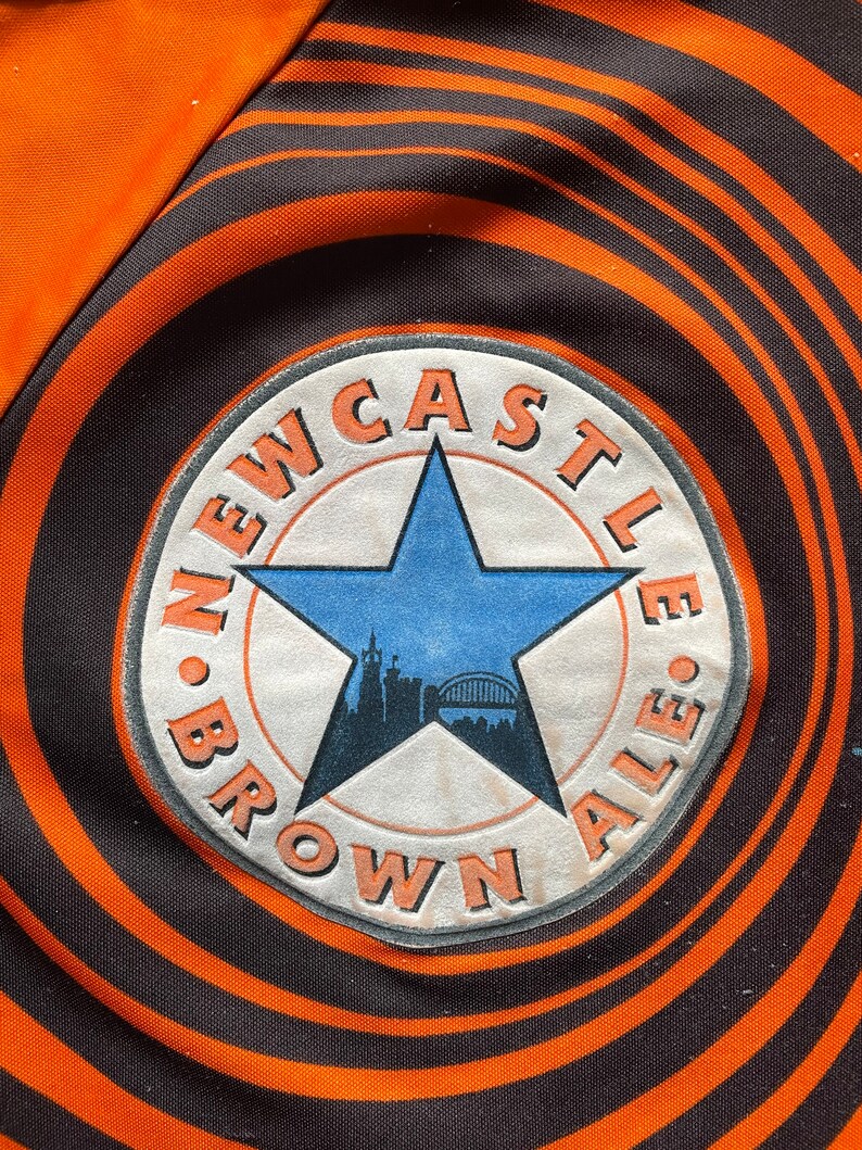 May include: A black and orange circular patch with the text "NEWCASTLE BROWN ALE" in a white circle with a blue star and a cityscape inside the star.