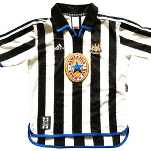 May include: A white and black striped soccer jersey with blue accents. The jersey features the Adidas logo, a team crest, and a patch with text. The collar and sleeves have blue trim.
