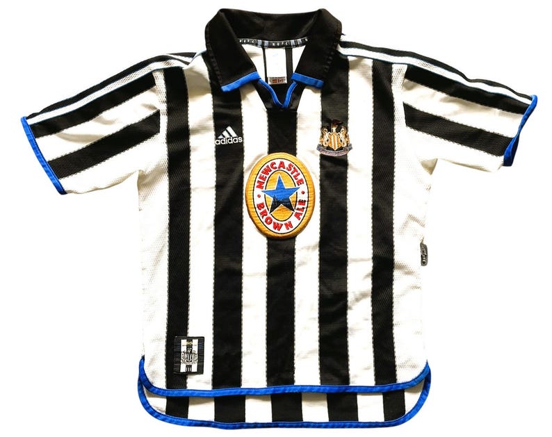 May include: A white and black striped soccer jersey with blue accents. The jersey features the Adidas logo, a team crest, and a patch with text. The collar and sleeves have blue trim.