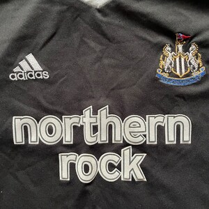 May include: Black football shirt with a white Adidas logo, a Newcastle United crest, and the words "northern rock" in white.