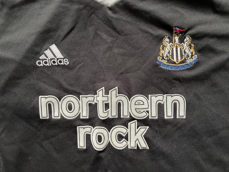 May include: Black football shirt with a white Adidas logo, a Newcastle United crest, and the words "northern rock" in white.