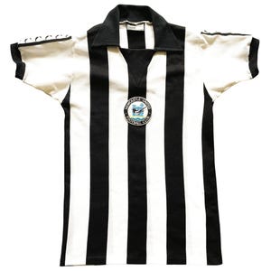 Może przedstawiać: Koszulka Newcastle United Football Club w stylu vintage. Koszulka ma czarno-białe pionowe paski, czarny kołnierz i kr&oacute;tkie rękawy. Na piersi znajduje się okrągła odznaka z nazwą i logo klubu.