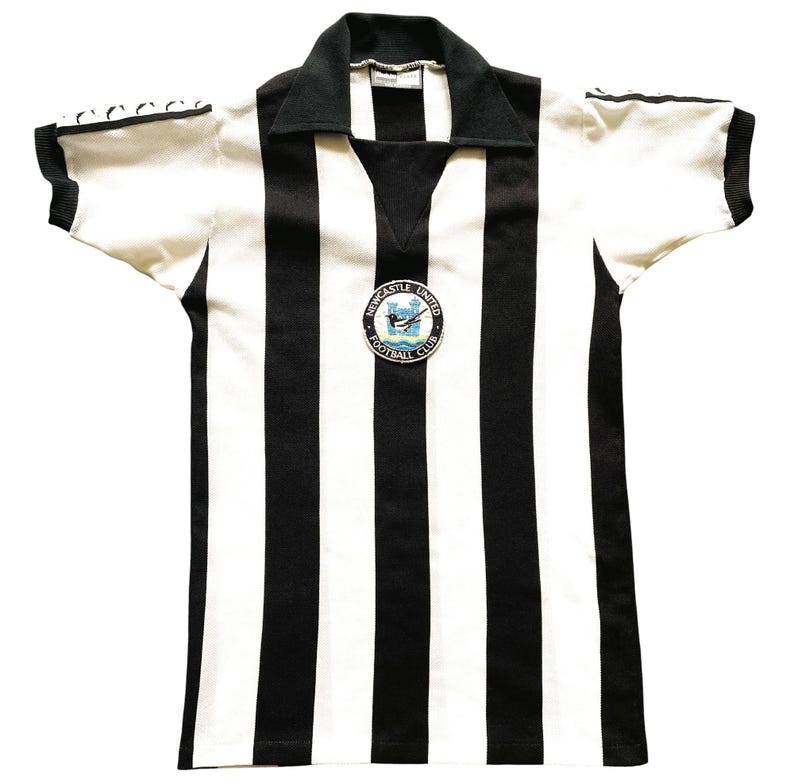 Może przedstawiać: Koszulka Newcastle United Football Club w stylu vintage. Koszulka ma czarno-białe pionowe paski, czarny kołnierz i kr&oacute;tkie rękawy. Na piersi znajduje się okrągła odznaka z nazwą i logo klubu.