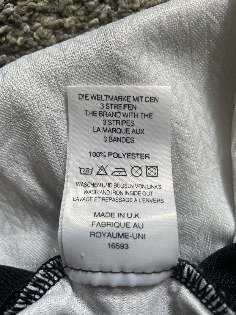 May include: White tag with black text that reads "Die Weltmarke mit den 3 Streifen", "The Brand with the 3 Stripes", "La Marque aux 3 Bandes", "100% Polyester", "Waschen und B&uuml;geln von Links", "Wash and Iron Inside Out", "Lavage et Repassage a l'Envers", "Made in U.K.", "Fabrique au Royaume-Uni", "16593". The tag also has a 40 degree Celsius washing symbol, a do not bleach symbol, a tumble dry low symbol, and an iron low symbol.