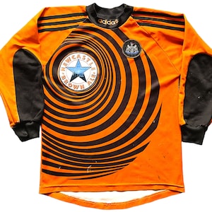 May include: Orange and black long-sleeved soccer jersey with a black and white swirl pattern. The jersey has a black star and the text "Newcastle Brown Ale" on the front. The Adidas logo is on the chest and the Newcastle United crest is on the right shoulder.