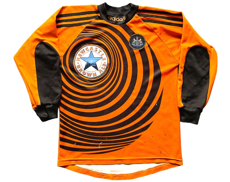 May include: Orange and black long-sleeved soccer jersey with a black and white swirl pattern. The jersey has a black star and the text "Newcastle Brown Ale" on the front. The Adidas logo is on the chest and the Newcastle United crest is on the right shoulder.