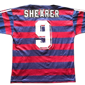 May include: A red and blue striped jersey with the name "SHEARER" and the number "9" in white. The jersey has white stripes on the sleeves.
