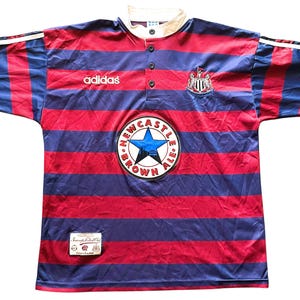 May include: A vintage Newcastle United soccer jersey. The shirt features red and blue horizontal stripes, a white collar, and the Adidas logo. It has a round patch with a blue star and the words "Newcastle Brown Ale".