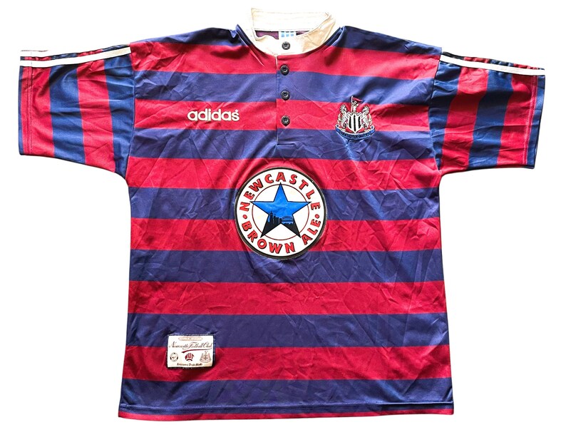 May include: A vintage Newcastle United soccer jersey. The shirt features red and blue horizontal stripes, a white collar, and the Adidas logo. It has a round patch with a blue star and the words "Newcastle Brown Ale".