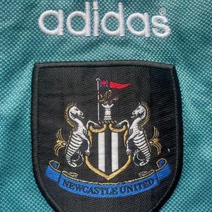 May include: A black and white embroidered patch with the Newcastle United crest. The crest features two white seahorses flanking a shield with black and white stripes. A blue ribbon with the words "Newcastle United" is below the shield. The patch is on a teal background with the word "adidas" in white above it.