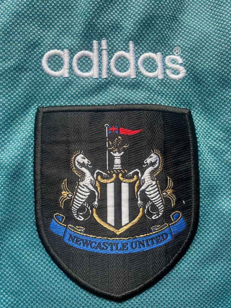 May include: A black and white embroidered patch with the Newcastle United crest. The crest features two white seahorses flanking a shield with black and white stripes. A blue ribbon with the words "Newcastle United" is below the shield. The patch is on a teal background with the word "adidas" in white above it.