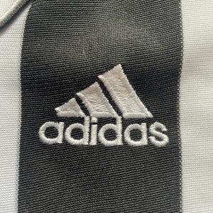 May include: Close-up of a black and white striped fabric with the Adidas logo embroidered in white. The Adidas logo is a white trefoil with the word "adidas" stitched below it.